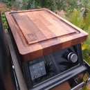 The BBQ Boards® Traeger Ironwood XL Pellet Bin Cutting Board sits atop a grill control panel, featuring the BBQ Boards® logo in one corner, with greenery and purple flowers in the background.