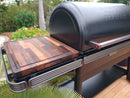 A Traeger grill with the BBQ Boards® Traeger Ironwood XL Side Cutting Board on its side shelf stands outdoors, surrounded by bright green plants and colorful flowers for a vibrant grilling setup.