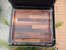 The BBQ Boards® Traeger Ironwood XL Side Cutting Board features a grooved edge and stamped logo, serving as an ideal grill prep surface outdoors on the Ironwood XL side board among plants and paving stones.