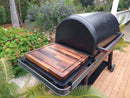 A BBQ Boards® Traeger Ironwood XL Side Cutting Board from BBQ Boards® sits on a Traeger grill’s side shelf on a brick patio, surrounded by lush greenery and flowers, with trees and a white railing in the background.