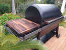 A large black Traeger grill on a patio showcases the BBQ Boards® Traeger Ironwood XL Side Cutting Board as its prep surface, with green foliage and colorful flowers in the vibrant background.