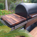 The BBQ Boards® Traeger Ironwood XL Side Cutting Board by BBQ Boards® is shown on the side board of a black outdoor grill, surrounded by vibrant garden greenery, flowers, and trees.
