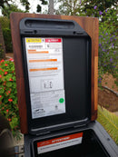 Close-up of a BBQ Boards® Traeger Ironwood Pellet Bin Cutting Board by BBQ Boards®, placed on a garden BBQ prep surface with greenery, flowers, and the Traeger Ironwood pellet grill lid displaying caution labels in the background.