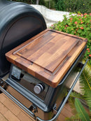 A BBQ Boards® Traeger Ironwood Pellet Bin Cutting Board sits on the side shelf of a Traeger Ironwood pellet grill with control dials, surrounded by plants and flowers against a white wall for a fresh outdoor vibe.