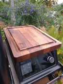 A BBQ Boards® Traeger Ironwood Pellet Bin Cutting Board sits on a Traeger Ironwood pellet grill outdoors, surrounded by greenery and purple flowers.