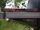 The BBQ Boards® Traeger Ironwood Side Cutting Board with a handle slot sits on a textured metal grill prep workspace, while green foliage and trees appear in the background.