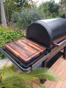 The BBQ Boards® Traeger Ironwood Side Cutting Board by BBQ Boards® creates an ideal grill prep space on your outdoor grill, set on a patio amid lush greenery and blooming flowers.