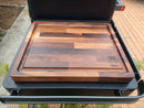 The BBQ Boards® Traeger Ironwood Side Cutting Board, featuring a groove and varied wood tones with the BBQ Boards® logo, rests on a black grill as a perfect outdoor prep space. Bricks and plants can be seen in the background.