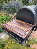 The BBQ Boards® Traeger Ironwood Side Cutting Board rests on a black grill’s side board, surrounded by green plants, purple flowers, and a garden path to create the perfect grill prep space.