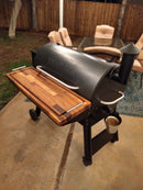 A Traeger smoker grill with a BBQ Boards® Traeger Pro 34 Front Cutting Board is on a patio beside a glass outdoor table and chairs. A metal bucket hangs from the grill, and a wooden fence stands in the background.