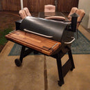 A Traeger smoker grill with a BBQ Boards® Traeger Pro 34 Front Cutting Board is on the patio. Behind it, a glass table and four cushioned chairs are on a green rug near a wooden fence.