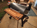 A BBQ Boards® Traeger Pro 34 Front & Pellet Bin Cutting Boards (Pair) are on a Traeger wood pellet grill on a patio. Seasoning containers are on the grill, with patio chairs, a table in the background, and a small metal bucket hanging on the side.