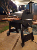 A black pellet grill with a wooden side shelf features BBQ Boards® Traeger Pro 34 Front & Pellet Bin Cutting Boards (Pair) on a covered patio at night. A metal bucket hangs on the side, with trees and a fence in the background.