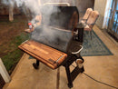 A pellet grill with smoke rising stands on a patio, featuring BBQ Boards® Traeger Pro 34 Front & Pellet Bin Cutting Boards (Pair) for easy prep. A metal bucket hangs at the side, with patio chairs and a table in the background.