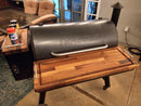 The BBQ Boards® Traeger Pro 34 Front & Pellet Bin Cutting Boards (Pair) are mounted on this wood pellet grill, with seasoning containers on the side shelf and patio furniture in the background.