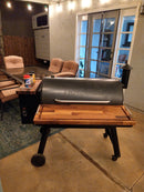 A Traeger wood pellet grill with BBQ Boards® Traeger Pro 34 Front & Pellet Bin Cutting Boards is on a concrete patio, surrounded by cushioned patio chairs, a table, string lights, and a sliding glass door in the background.
