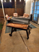 A Traeger Pro 34 grill with BBQ Boards® Traeger Pro 34 Front & Pellet Bin Cutting Boards (Pair) sits on a patio at night, near outdoor chairs and a table with grilling supplies. String lights hang above, with a wooden fence and house behind.