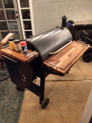 A black Traeger pellet grill features BBQ Boards® Traeger Pro 34 Front & Pellet Bin Cutting Boards (Pair), adding shelf space for spice containers. The setup is on a patio by a glass door and an outdoor rug.