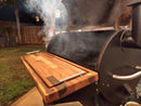 A smoky outdoor grill with the lid open, smoke rising, is set on a patio beside BBQ Boards® Traeger Pro 34 Front & Pellet Bin Cutting Boards (Pair) by BBQ Boards®, with a grassy yard and wooden fence at night.