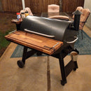 A pair of BBQ Boards® Traeger Pro 34 Front & Pellet Bin Cutting Boards accents this outdoor pellet grill, featuring a wooden side shelf and metal bucket, displayed on a patio with cushioned chairs, table, and a fence in the background.