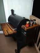 On a deck near a white wall, BBQ Boards® Traeger Pro 780 Front & Pellet Bin Cutting Boards (Pair) by BBQ Boards® are shown installed on a black Traeger grill, serving as prep shelves with BBQ sauce and spices placed on the right board.
