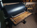 A pair of BBQ Boards® Traeger Pro 780 Front & Pellet Bin Cutting Boards sits on a Traeger grill at night, with seasoning containers on the railing, a metal bucket hanging from the side, and bright lighting for easy grilling.