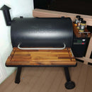 A Traeger wood pellet grill on a deck at night features BBQ Boards® Traeger Pro 780 Front & Pellet Bin Cutting Boards (Pair), with three spice containers on a side shelf and a wall light mounted nearby.