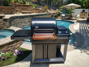 A BBQ Boards® Traeger Timberline Front Cutting Board is attached to a large black outdoor grill on a patio by a pool, with stone walls, flowers, lounge chairs, an umbrella, and greenery visible in the sunny background.