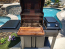 A BBQ Boards® Traeger Timberline Front Cutting Board sits atop a modern outdoor grill by the pool, surrounded by landscaped stone and plants on a sunny day. The grill lid is open, displaying clean metal grates.
