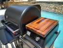The BBQ Boards® Traeger Timberline Pellet Bin Cutting Board by BBQ Boards® offers a spacious prep surface with an attached wooden board, shown beside a pool with a stone wall backdrop on a sunny day.