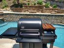 A black outdoor grill with a closed lid stands by a pool, featuring the BBQ Boards® Traeger Timberline Pellet Bin Cutting Board for extra prep space, with a control panel on the right. A stone wall and landscaped greenery complete the backdrop.