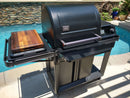 A black Traeger grill with a BBQ Boards® Traeger Timberline Side Cutting Board by BBQ Boards® on its side shelf sits on a concrete patio near a stone wall and plants, creating an ideal BBQ prep station by the pool.