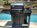 A BBQ Boards® Traeger Timberline Side Cutting Board by BBQ Boards® is attached to a black pellet grill on a patio next to a pool, stone retaining wall, and lush landscaping—ideal for sunny day BBQ prep.