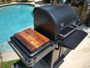 A black outdoor grill with a closed lid is by the pool. A BBQ Boards® Traeger Timberline Side Cutting Board from BBQ Boards® sits on the side shelf, creating a sunny, relaxed setting ideal for barbecues or cookouts.