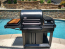 A black outdoor grill with a closed lid stands beside the BBQ Boards® Traeger Timberline Side Cutting Board, near a pool with stone walls and landscaping in the background.