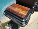 The BBQ Boards® Traeger Timberline Side Cutting Board sits on a metal grill shelf by a pool, surrounded by flowers and greenery. Sunlight highlights the wood grain and engraved BBQ Boards® logo on this premium prep station accessory.