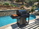 A BBQ Boards® Traeger Timberline Side Cutting Board sits beside a black outdoor grill and swimming pool, surrounded by stone walls, plants, and patio furniture in a sunny backyard.