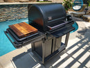 A black grill with a closed lid stands by a pool, featuring the BBQ Boards® Traeger Timberline Side Cutting Board on its side shelf—ideal for BBQ prep. Patio furniture and a hanging chair appear in the background.