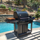 A BBQ Boards® Traeger Timberline Side Cutting Board from BBQ Boards® sits on a large black outdoor grill with a closed lid by the pool on a sunny patio, surrounded by greenery, stonework, and a tiered garden—an ideal BBQ prep station.
