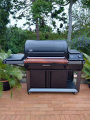 Spacious black outdoor smoker grill with BBQ Boards® Traeger Timberline XL Front Cutting Board, metal cabinets below, and right-side control panel—ideal for prepping and serving BBQ on the patio amid lush greenery.
