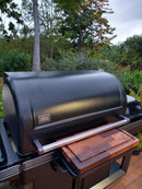 A large black Traeger grill with a closed lid sits on a garden patio, featuring a BBQ Boards® Traeger Timberline XL Front Cutting Board to add convenient prep space to the side shelf, surrounded by lush greenery.