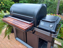 A modern outdoor grill features the BBQ Boards® Traeger Timberline XL Front Cutting Board by BBQ Boards®, a digital control panel, and spacious storage, all set on a brick patio with lush plants and colorful flowers.