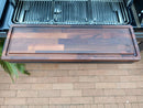 The BBQ Boards® Traeger Timberline XL Front Cutting Board by BBQ Boards® rests on a brick patio in front of a stainless steel grill, providing an ideal BBQ prep space. A small plant appears on the left side of the image.