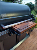 A Traeger grill with the BBQ Boards® Traeger Timberline XL Front Cutting Board sits on the patio, its pull-out side tray ready for prep. Surrounded by green plants and trees, it creates a lush, perfect BBQ space.
