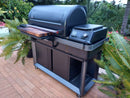 A sleek black outdoor grill with a side shelf, digital controls, and a BBQ Boards® Traeger Timberline XL Front Cutting Board sits on a brick patio surrounded by green plants and tropical foliage.