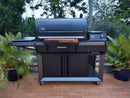 A BBQ Boards® Traeger Timberline XL Front Cutting Board by BBQ Boards® sits on a large black Broil King grill’s prep space on the patio, ready for your next cookout and surrounded by potted ferns and greenery.