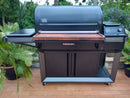 A large black outdoor pellet grill features the BBQ Boards® Traeger Timberline XL Front Cutting Board by BBQ Boards® as a BBQ prep space, with side shelves, control panel, and storage cabinet, standing on a patio among green plants.