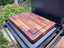 The BBQ Boards® Traeger Timberline XL Side Cutting Board rests atop a closed outdoor grill, offering an ideal prep space. Bright flowers and lush greenery add color to the background.