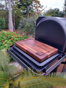 The BBQ Boards® Traeger Timberline XL Side Cutting Board by BBQ Boards® sits atop a closed outdoor grill, providing an ideal prep surface amid lush greenery and vibrant flowers in a beautiful garden setting.