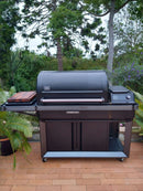 The BBQ Boards® Traeger Timberline XL Side Cutting Board by BBQ Boards® adds ample prep space to your grill, perfect for outdoor cooking on a brick patio surrounded by greenery and tall trees.